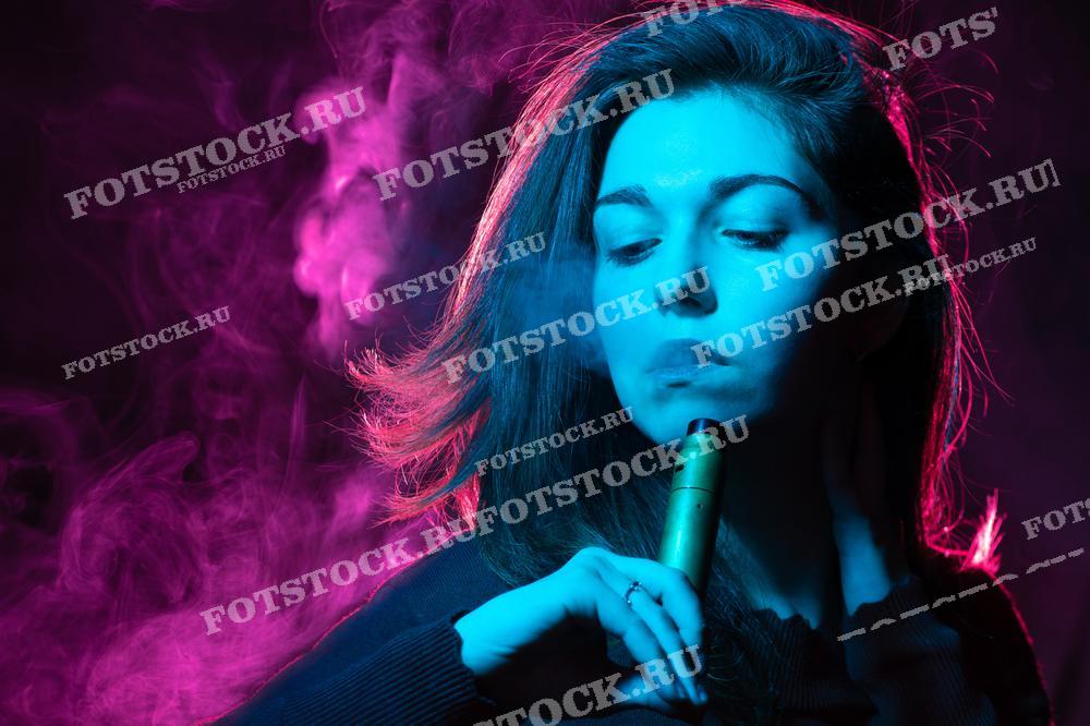 Фотография изображает молодую женщину, выпускающую пар от электронной сигареты. Лицо и фон освещены разноцветными огнями, создавая атмосферу ночного города и подчеркивая современные тренды вейпинга.